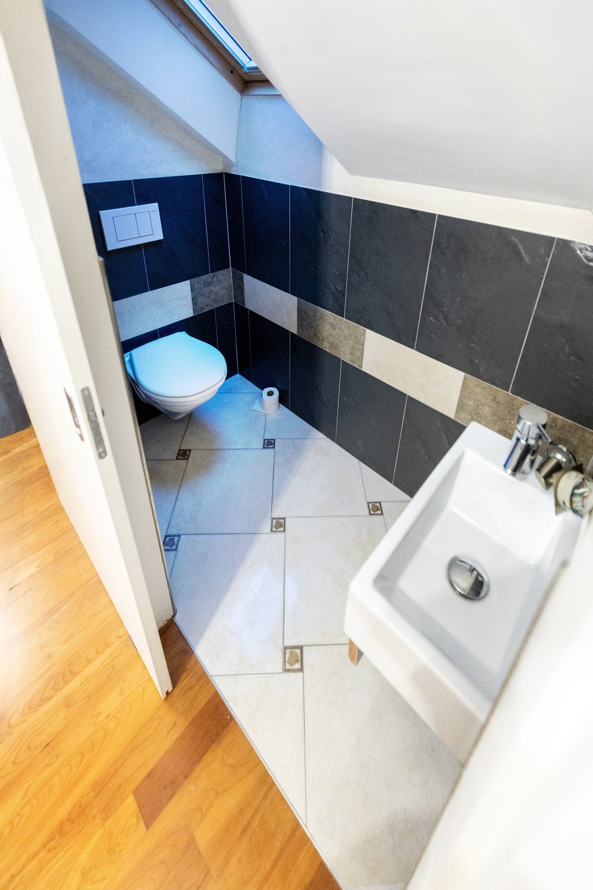 Modern bathroom with white toilet, dark and light blue tiled walls, white sink, wooden floor entry, and skylight window above
