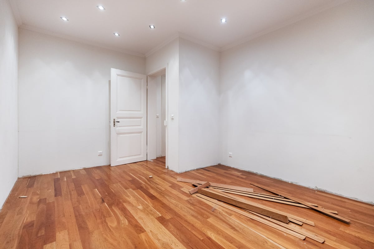 Empty room with white walls, recessed ceiling lights, white wooden door, and wooden flooring with scattered materials