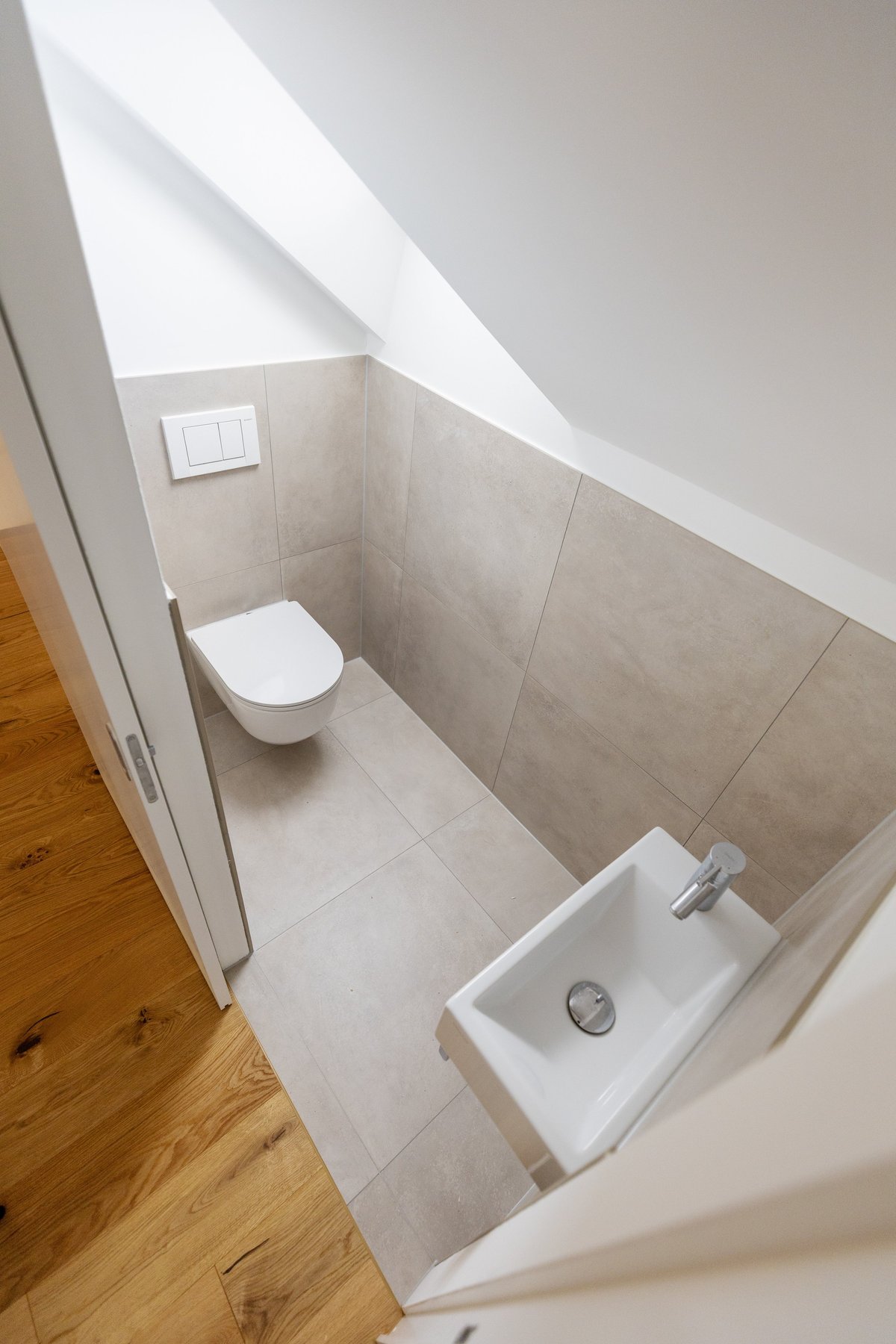Modern bathroom under sloped ceiling with white toilet and small sink on beige tile floor