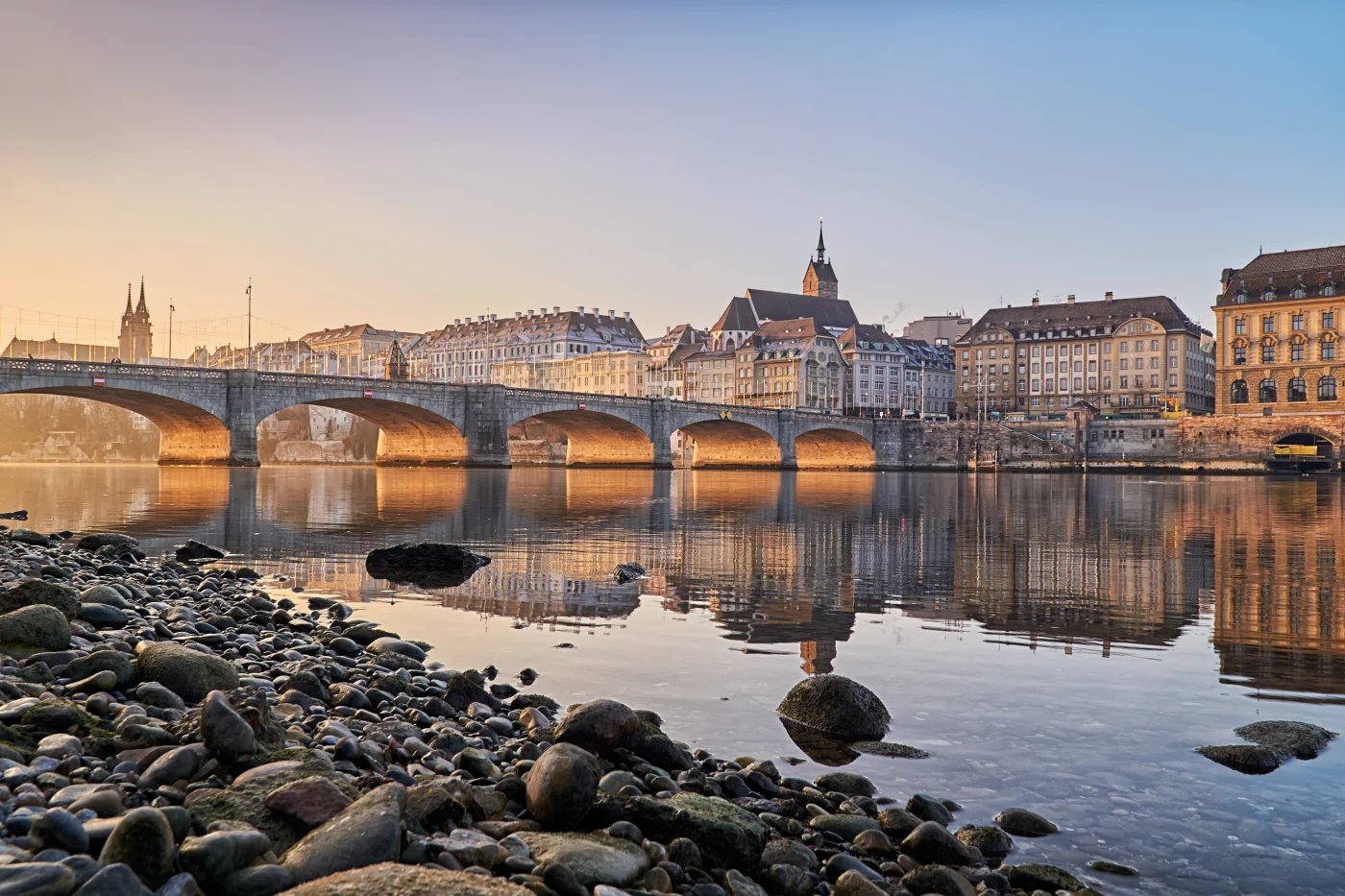 Historic stone bridge spanning calm river at sunrise with medieval city skyline, church spires, and classical architecture reflected in water